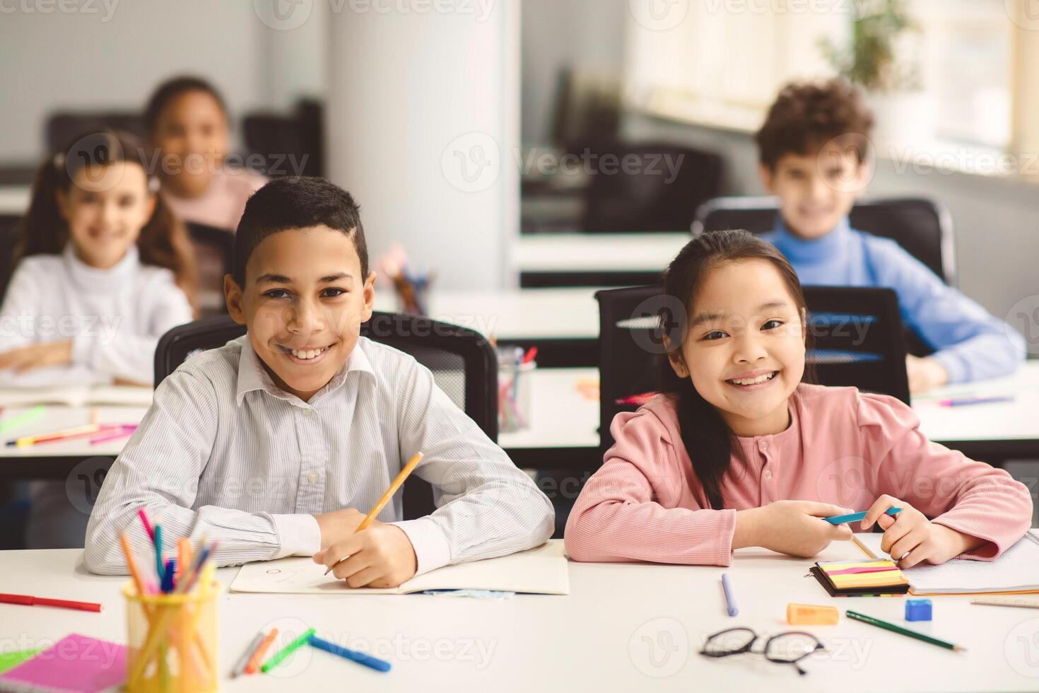 Education And Children Concept. Portrait of happy smiling diligent boy and girl sitting at table in classroom at elementary school with group of classmates in background and posing, looking at camera photo