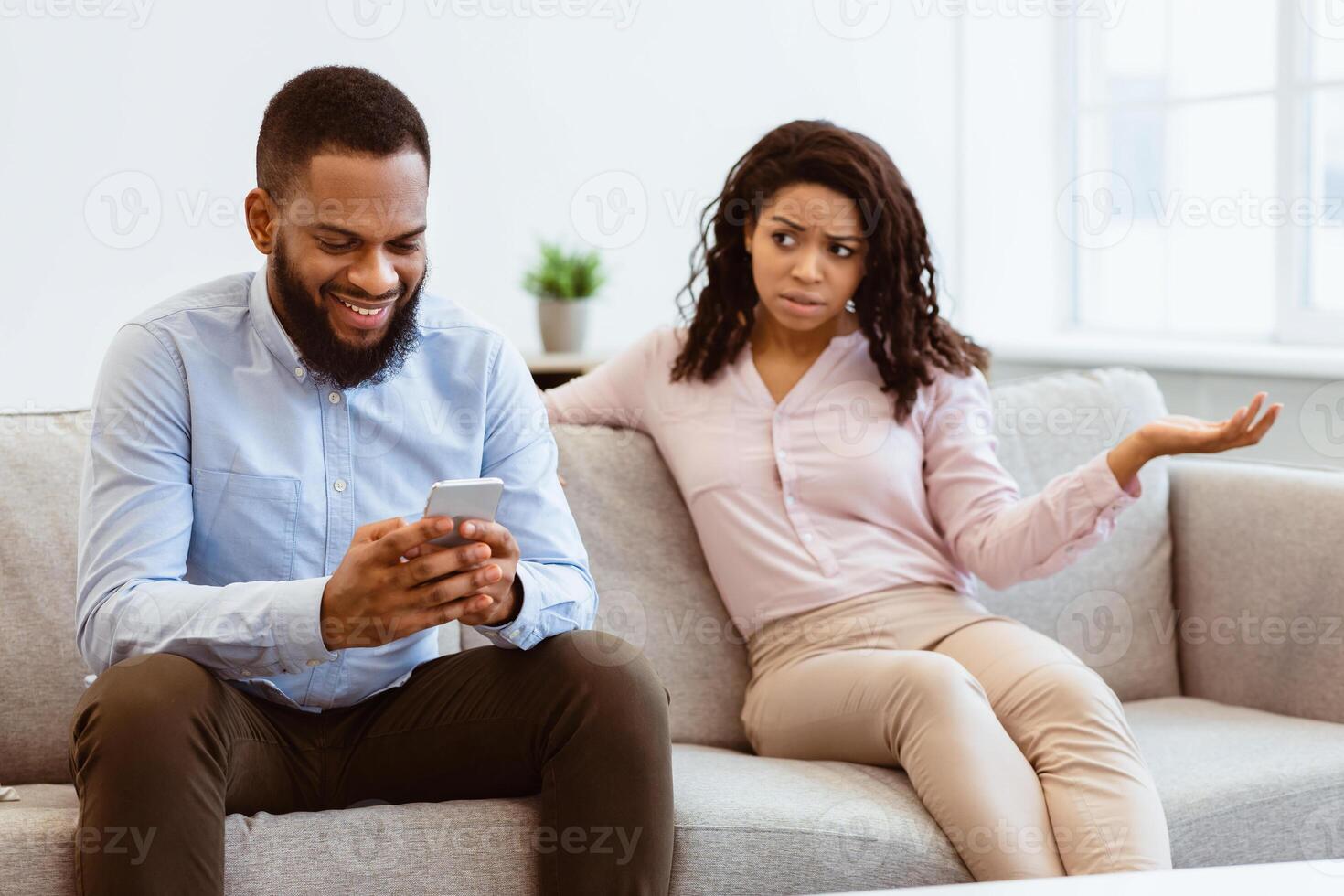 Phubbing. Angry young black woman looking at her boyfriend stuck in smartphone, unhappy about neglect and lack of communication. Jealous lady and gadget addicted man sitting on couch at home photo
