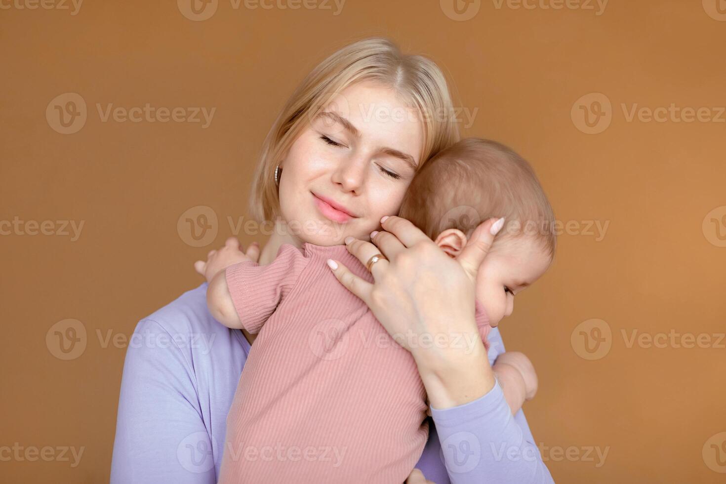 Happy motherhood concept. Loving young mother blonde woman cuddling with her cute little baby, brown studio background. Beautiful mom and sweet little kid toddler hugging and bonding photo