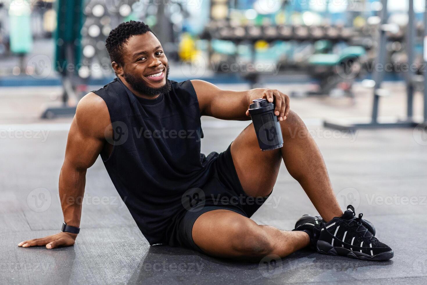 Happy african american bodybuilder holding mug with water or sports drink, sitting on floor at gym, resting, empty space. Smiling black muscular man having break while exercising at modern gym photo