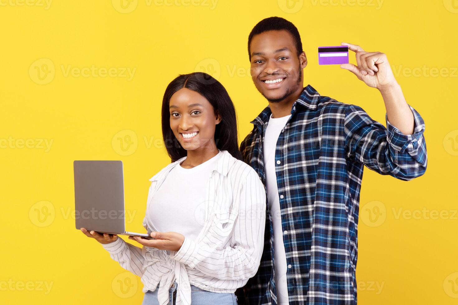 Quick home purchases on self-isolation during covid-19 quarantine. Smiling african american husband and wife hold laptop and show credit card to camera, isolated on yellow background, studio shot photo
