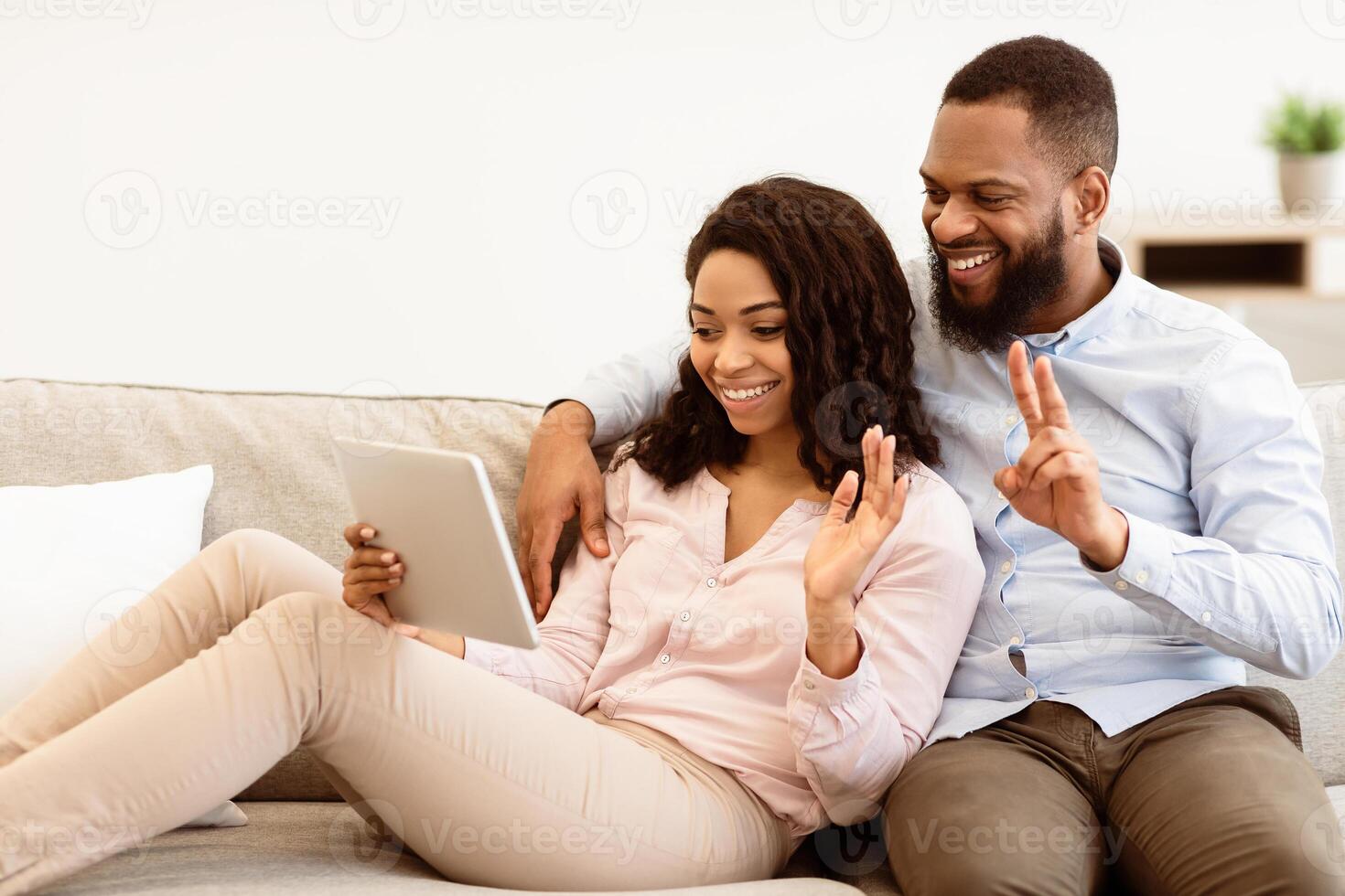 Virtual Communication. Happy black couple having online conference with friends or family using digital tablet, waving hand to web camera, sitting on the sofa in bedroom at home photo