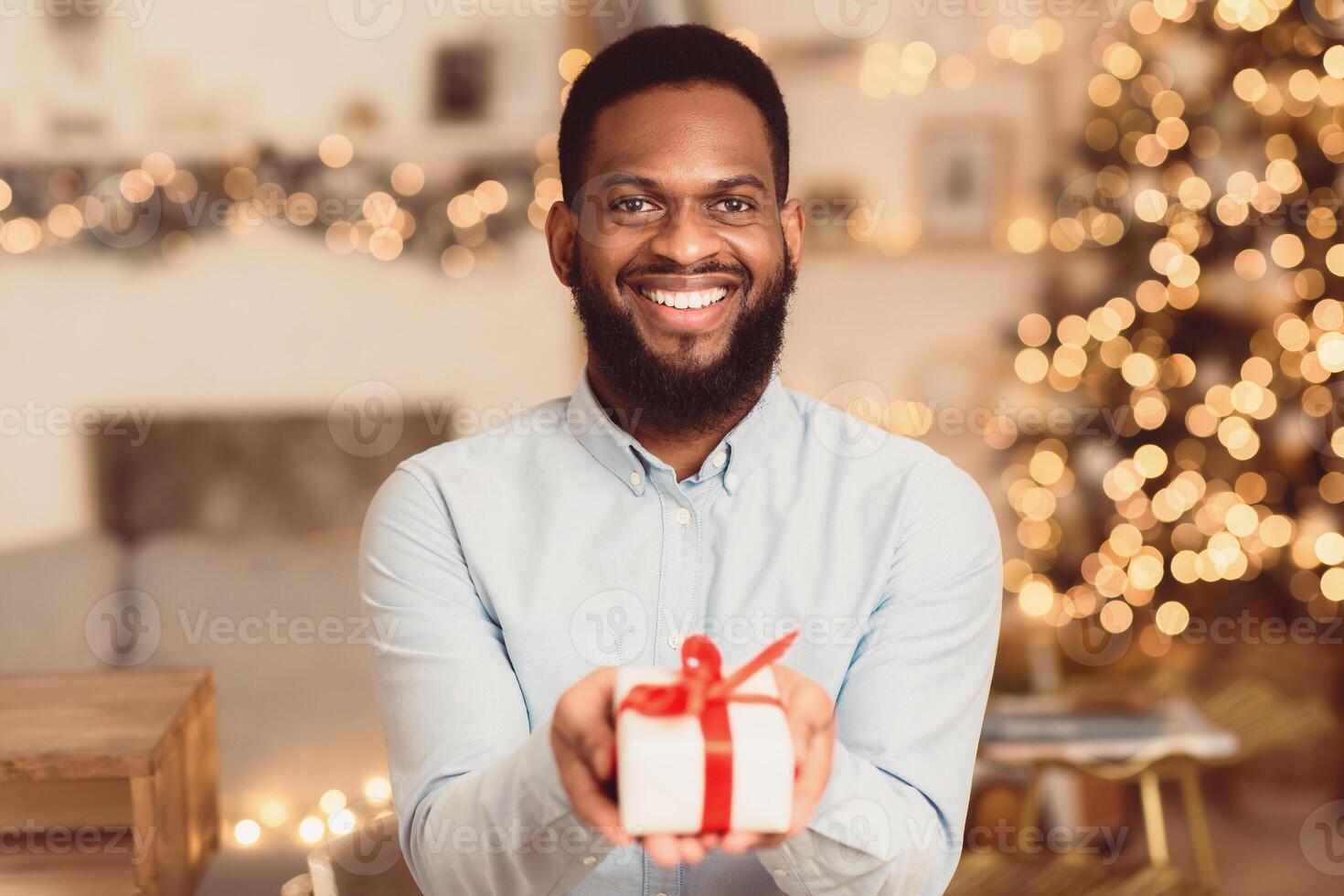 Celebration. Portrait of smiling handsome bearded african american man holding present gift box with red ribbon and showing it to camera, blurred background. Love, Bonding, Relationship Concept photo