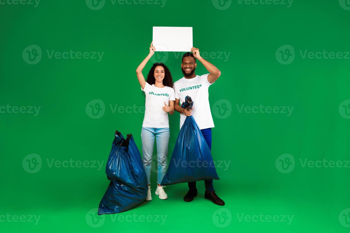 Two Diverse Volunteers Holding Blank Paper Board Posing With Plastic Bags Full Of Wasted Junk Standing Over Green Studio Background. Volunteering Work Concept. Mockup photo