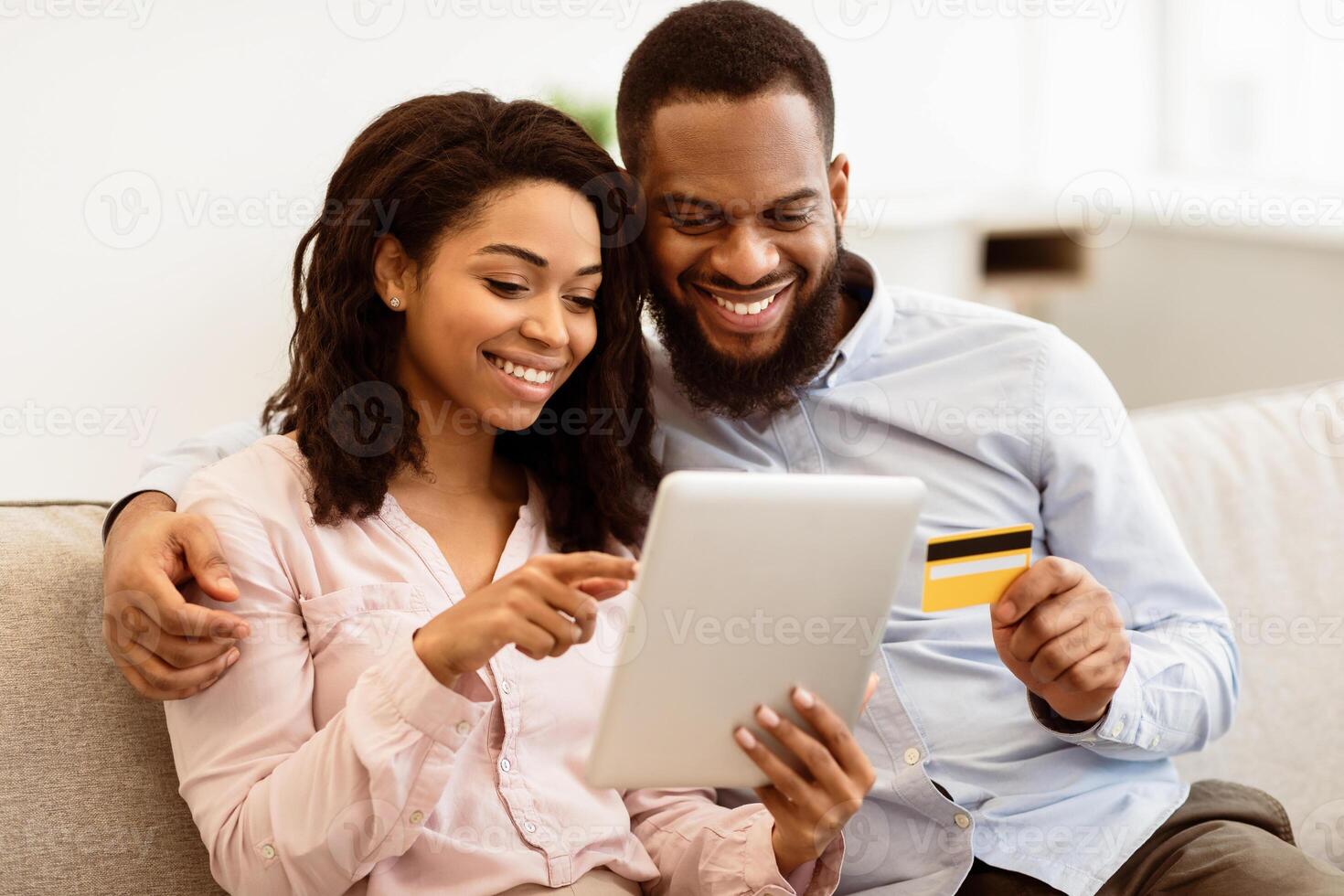Online Shopping And Ecommerce. Happy young african american family of two people sitting on sofa at home, choosing and buying clothes or products online, paying with credit card using digital tablet photo