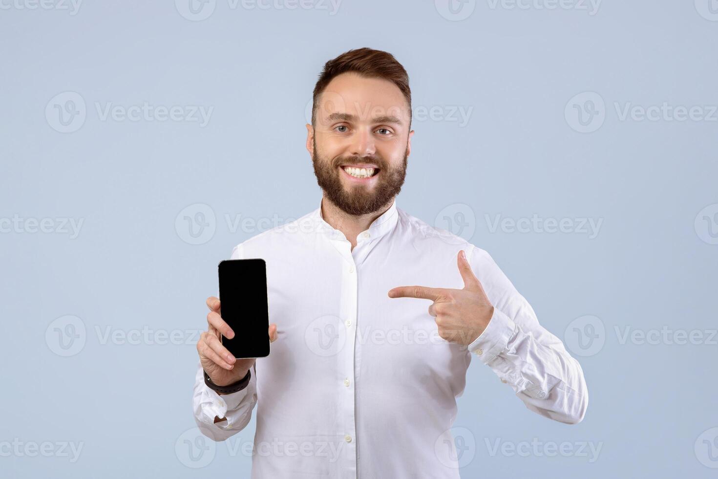 Happy bearded guy in formal wear pointing at smartphone with blank screen over grey studio background, free space. Young man showing mobile phone with mockup for website or app photo