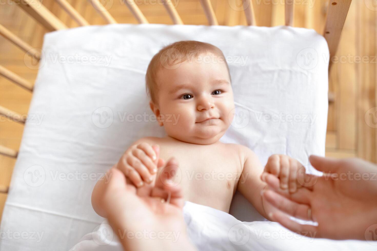 Cute little baby boy or girl laying in cot, holding mom's hands, ready to wake up. Top view of cute blonde toddler waking up, lying in wooden baby bed, grabbing mother fingers, copy space photo