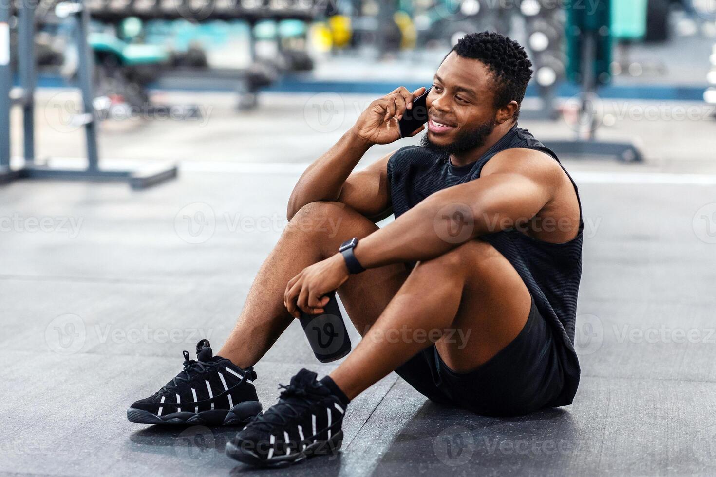 Muscular black guy sitting on floor in gym, holding bottle with protein or water and talking on smartphone, looking at copy space. Smiling african american athletic man resting after workout photo
