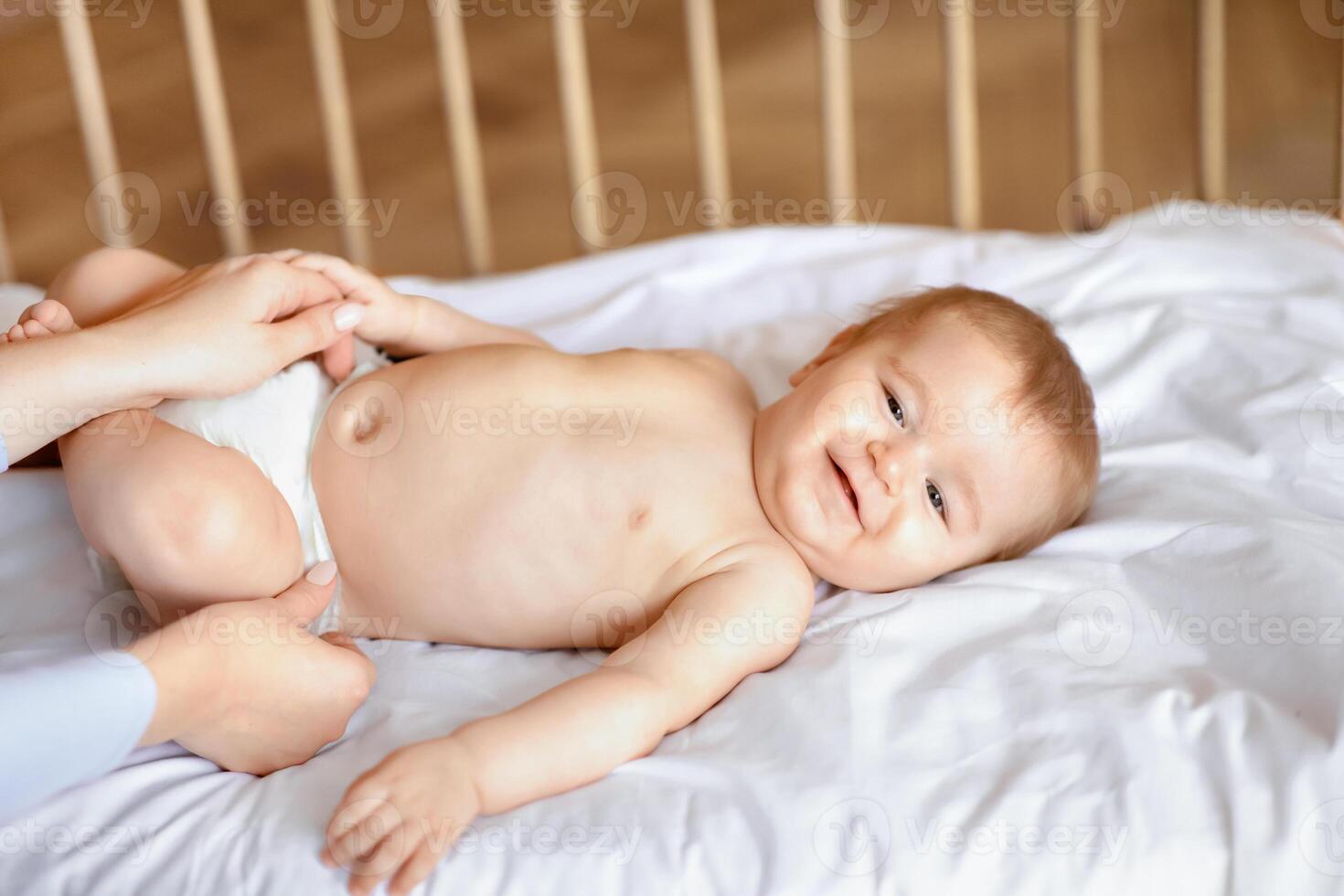 Adorable baby boy or girl laying in wooden bed, having fun while mom changing diaper, closeup. Smiling little kid laying in crib, enjoying morning after waking up, unrecognizable woman changing nappy photo
