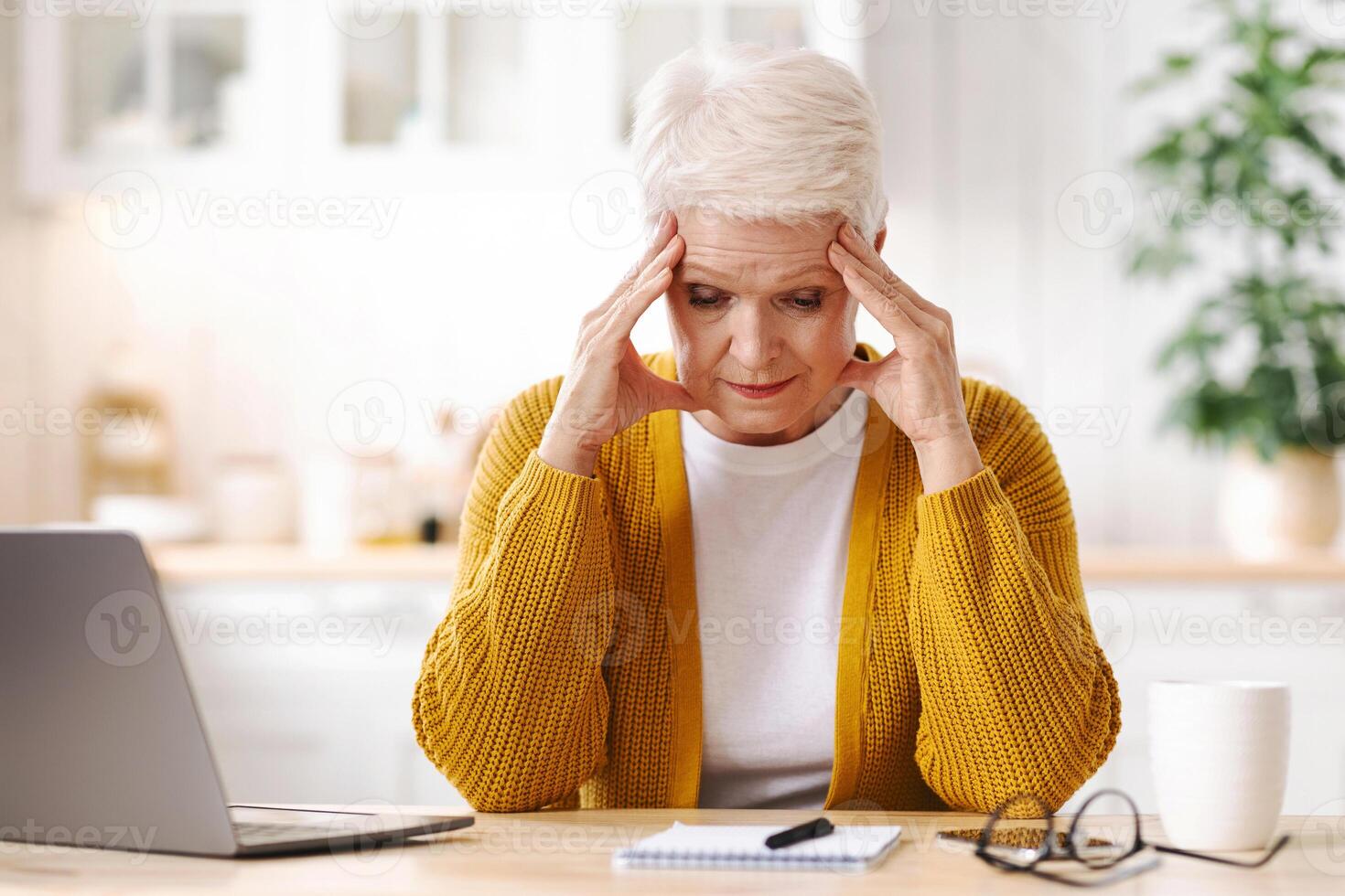 Exhausted mature grey-haired woman freelancer working from home, using laptop, sitting in kitchen, copy space. Stressful senior lady taking notes, sitting in front of computer, touching her head photo
