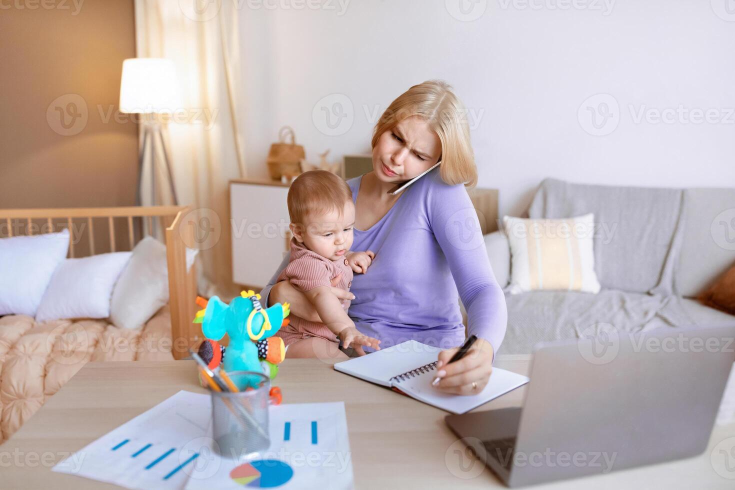 Busy young mother with little baby on her lap having business conversation on mobile phone, taking notes, sitting in front of laptop, home interior, copy space. Working mothers concept photo