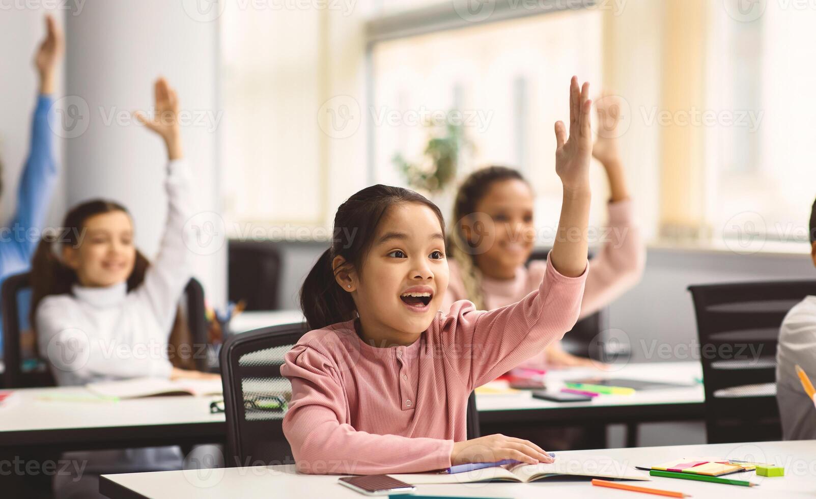 Education, Elementary School, Learning and People Concept. Diverse group of smart junior school kids sitting at desks in classroom and raising hands for an answer, studying with pleasure photo