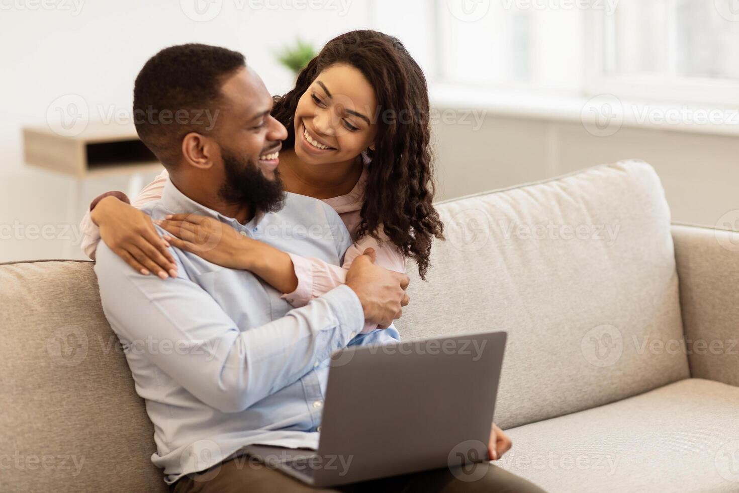 Happy Loving Family. Cheerful african american couple spending time together, looking at each other, man sitting on sofa at home, relaxing or working, positive wife hugging him from the back photo