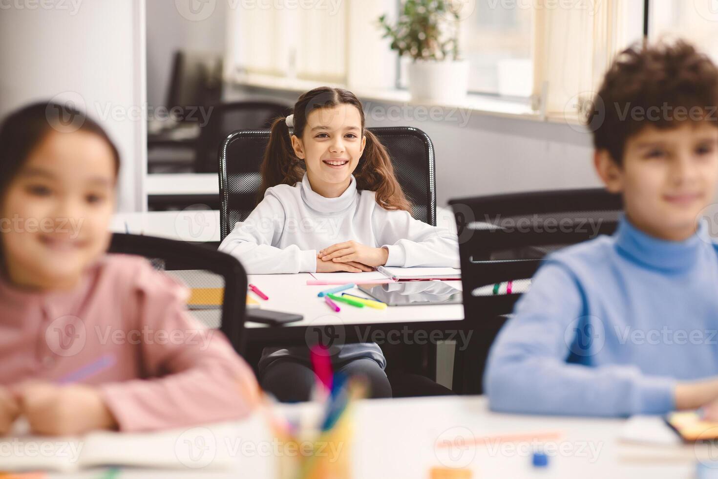 Elementary Education. Portrait of small girl with ponytails sitting at table in classroom at primary school or kindergarten with group of classmates. Return back to school after coronavirus quarantine photo