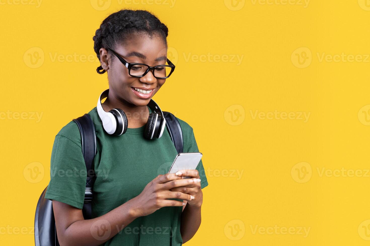 Happy African Female Student Using Smartphone Networking And Texting Online Standing Over Yellow Background. Young Woman Using Mobile Application On Phone. Studio Shot photo