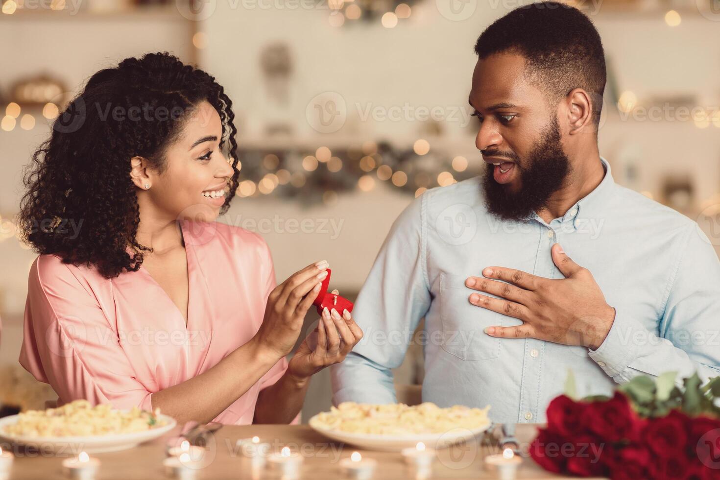 Be My Husband. Smiling black woman making proposal to her surprised man, showing present, holding open box with marriage ring, asking to get engaged during romantic dinner at home or cafe photo