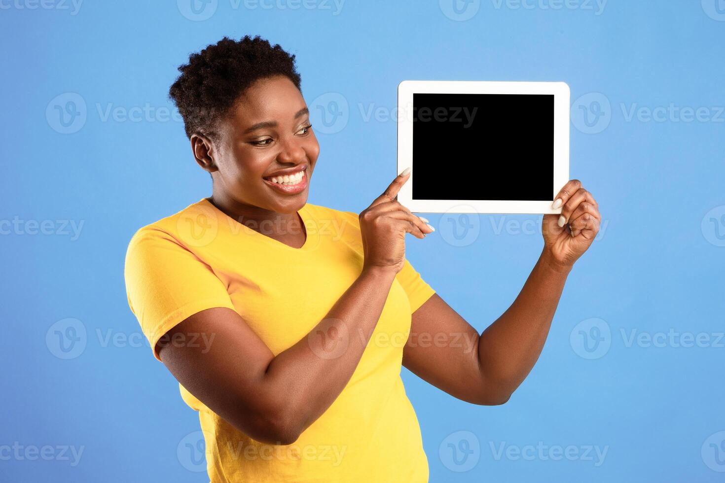 Smiling African American Woman Showing Digital Tablet Blank Screen Advertising Application Or Website Standing Over Blue Studio Background. Female Holding Computer With Empty Monitor. Mockup photo