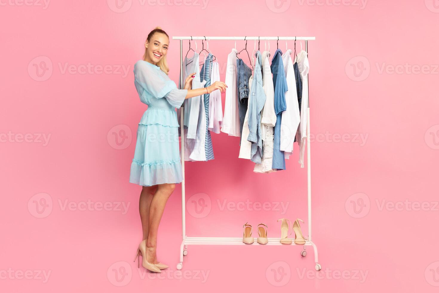 Full length vertical portrait of cheerful smiling woman standing at boutique wardrobe, choosing fashion stylish clothes at rack with hangers, looking at camera, looking for outfit, pink background photo