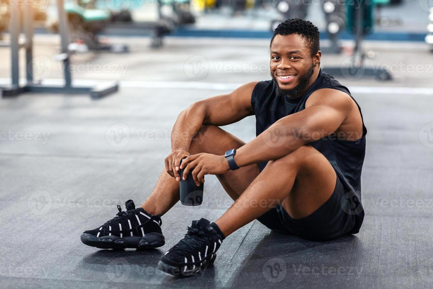 Smiling black guy in sportswear sitting on floor at gym, having protein drink, copy space. Cheerful african american sportsman having break during work out, holding bottle with water photo