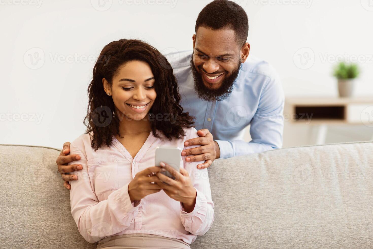 Cheerful millennial black couple browsing internet on smartphone, watching together on gadget. Handsome boyfriend embracing his girlfriend from back, lady sitting on the couch photo