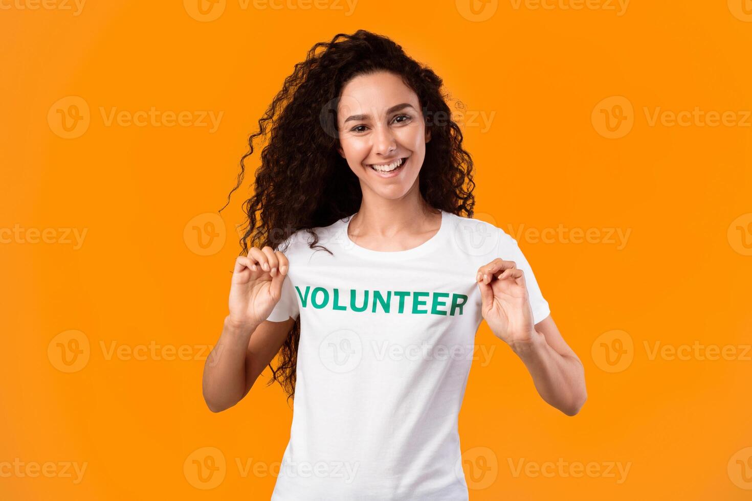 Proud activist woman showing volunteer text on her uniform t-shirt standing over yellow background, smiling to camera. Studio shot. Volunteering career and contribution concept photo
