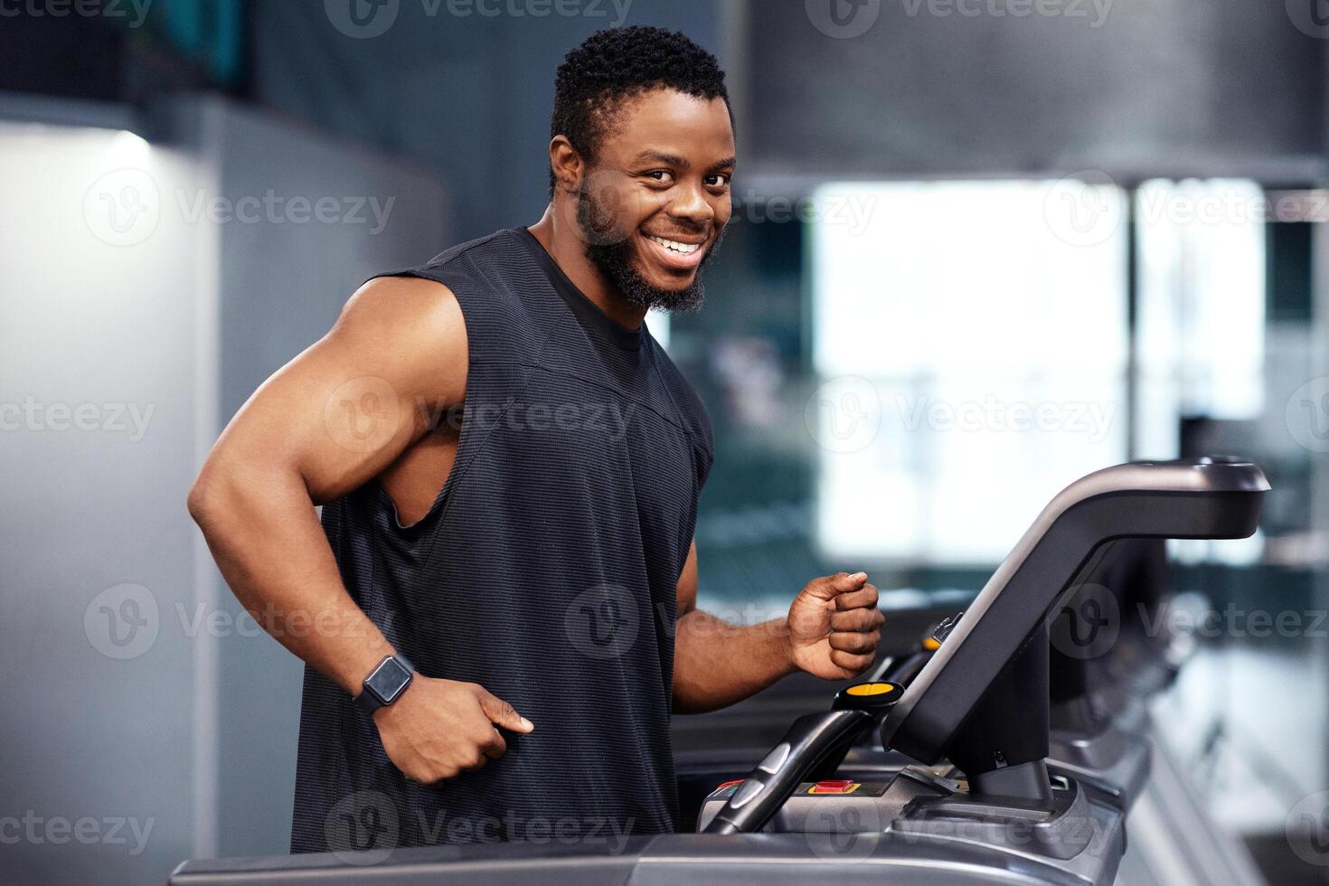 Jogging workout. Joyful black man in sportswear running on treadmill at gym, looking at camera and smiling, free space. Motivated african american guy in black sportswear spending time at gym, jogging photo