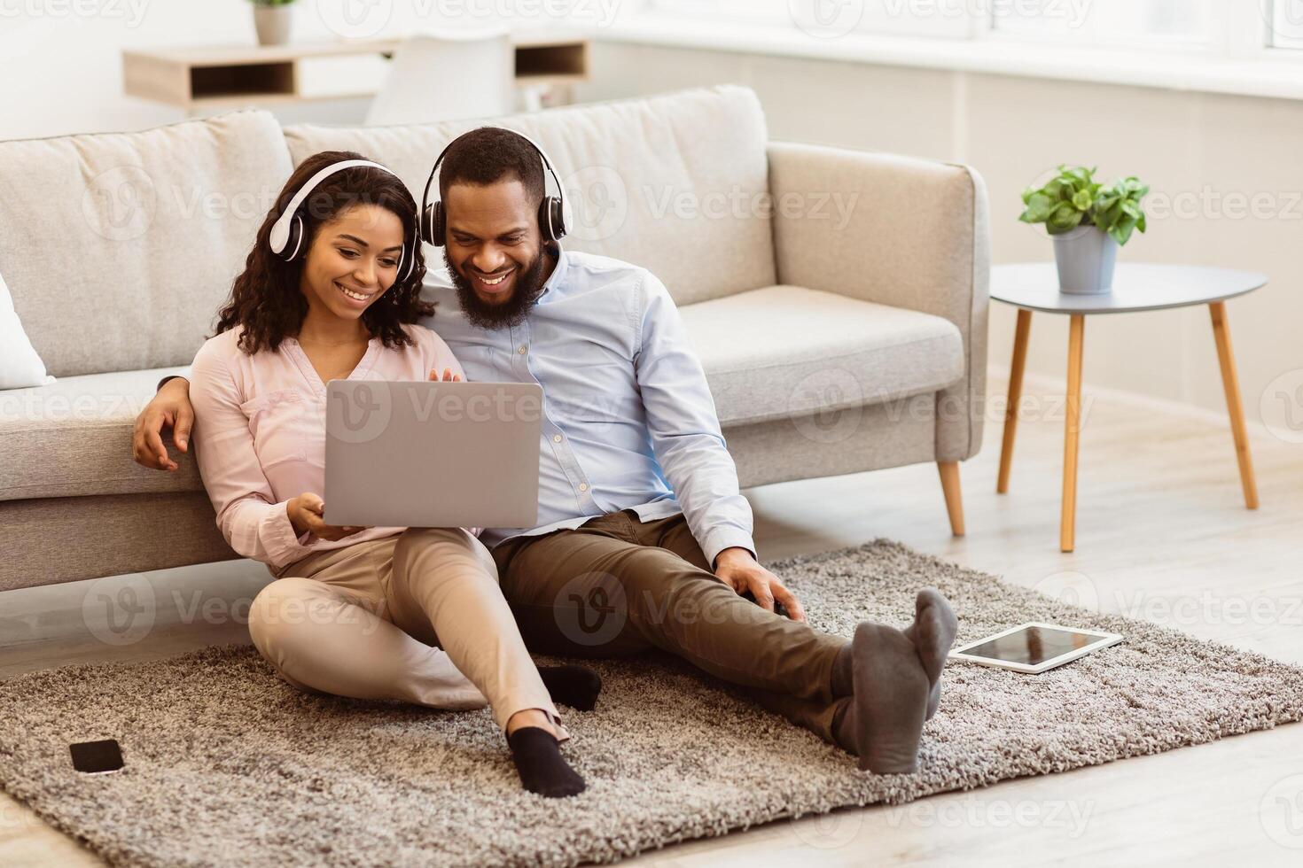 Virtual Communication. Happy black couple in headphones having online conference with friends or family using computer, waving hand to web camera, sitting on the floor carpet in bedroom at home photo