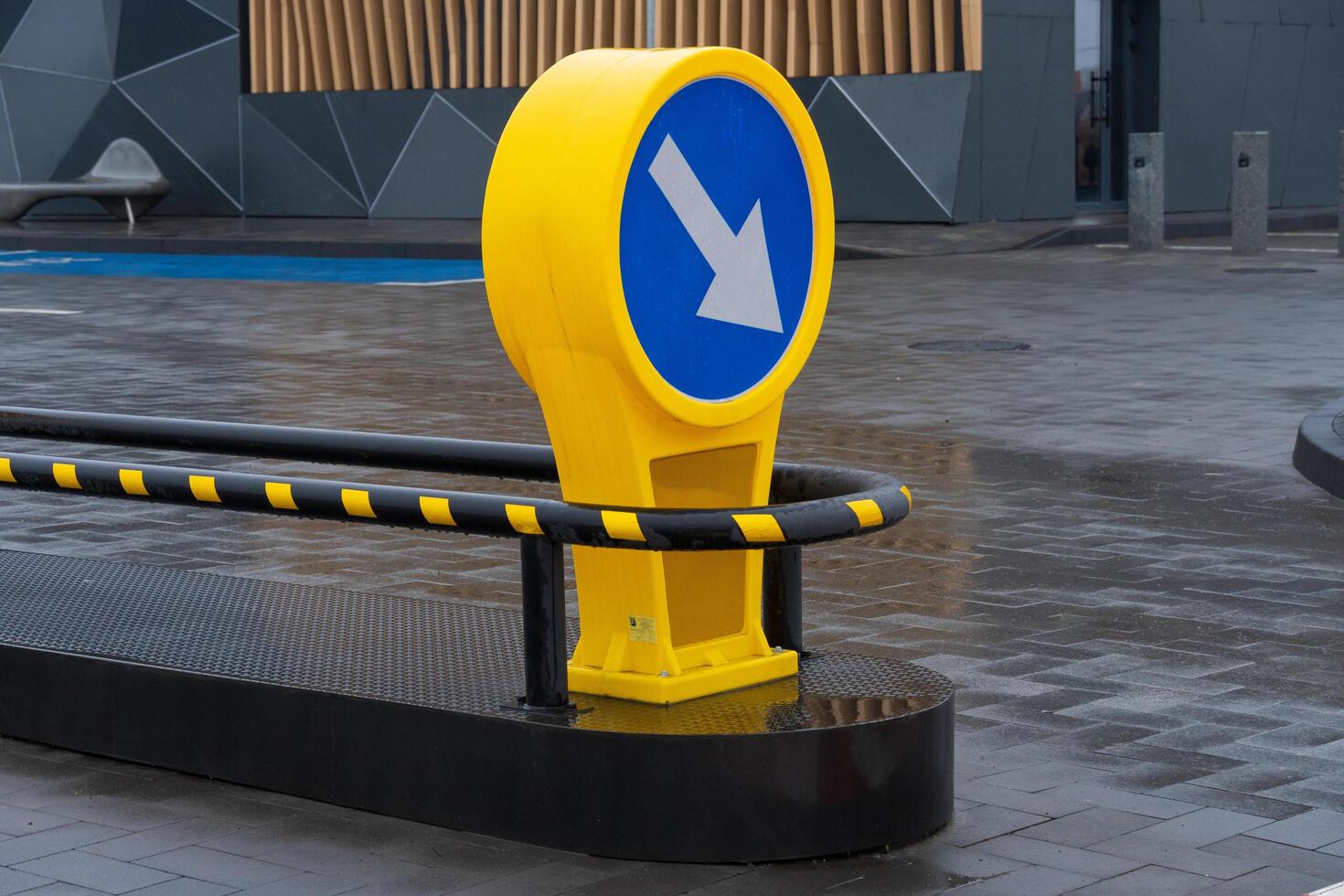 A bright yellow directional sign with an arrow points left at a contemporary city intersection. The wet pavement reflects the surroundings, highlighting the urban design and rainy weather photo
