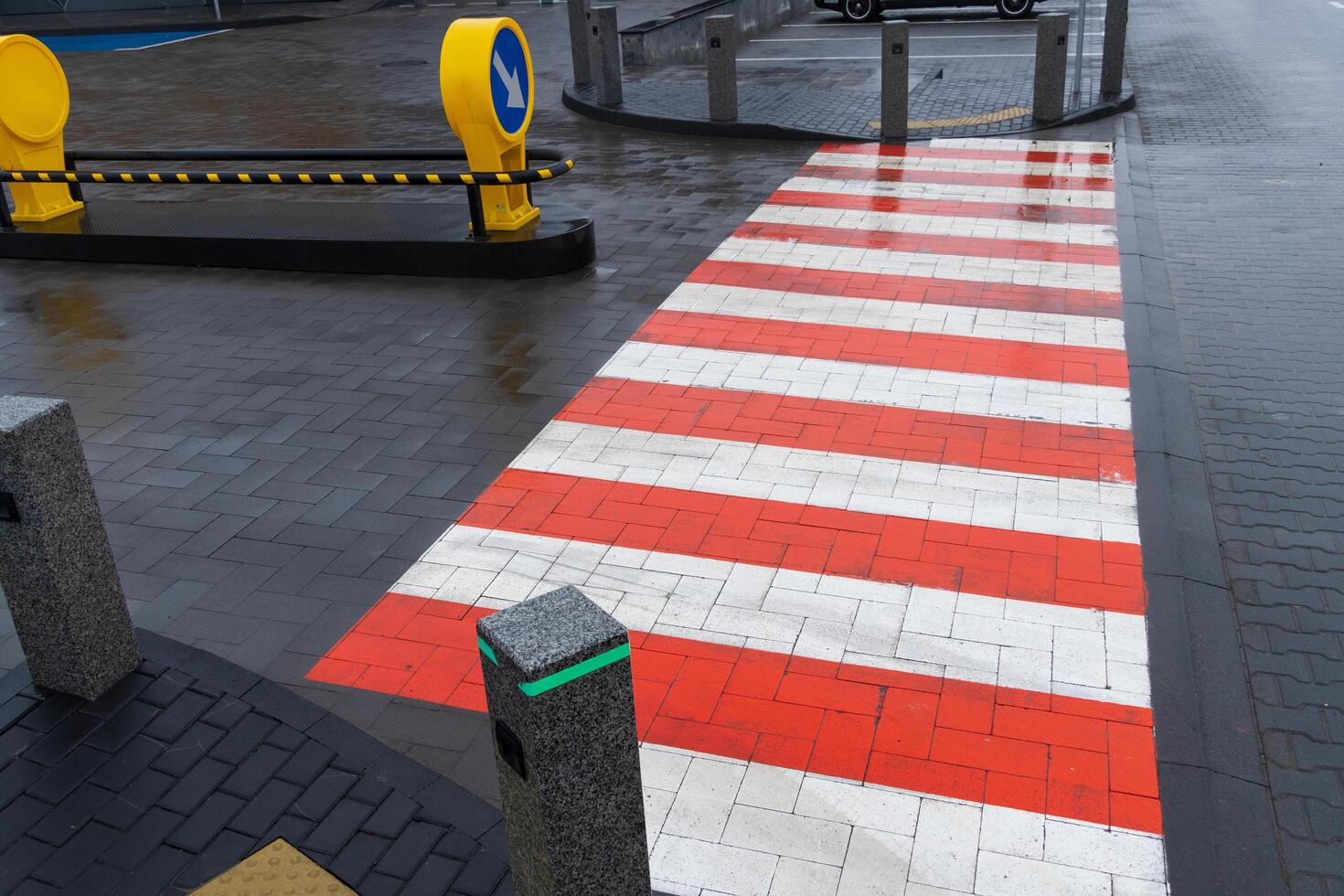 A vibrant red and white striped crosswalk stands out at an urban intersection, wet from recent rain. Traffic elements, including a roundabout sign, enhance the bustling city atmosphere photo