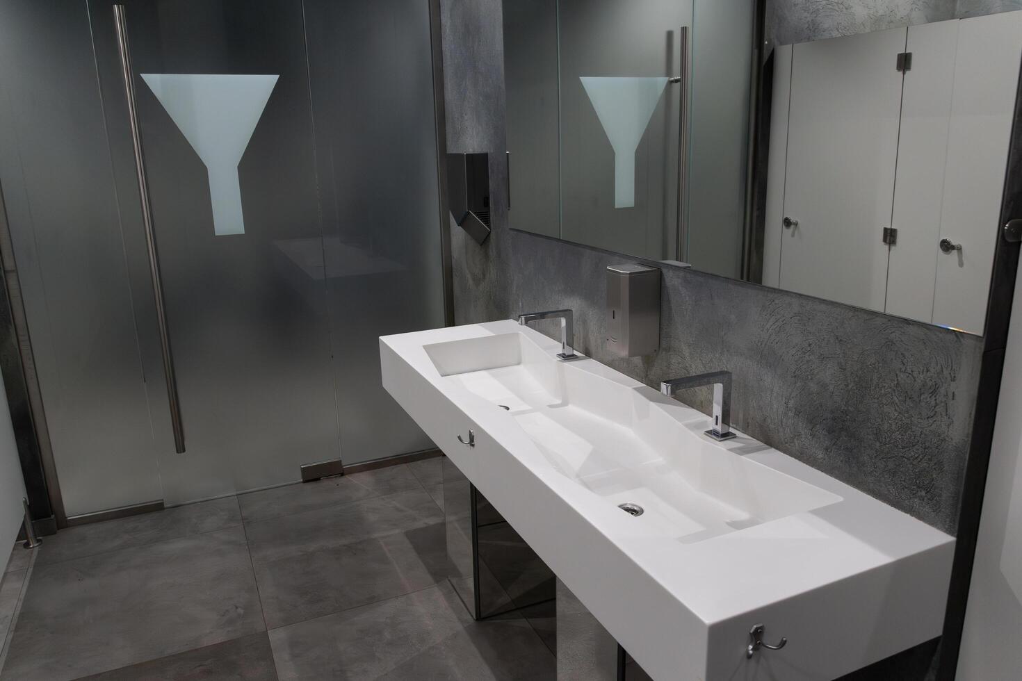 A spacious bathroom showcases a sleek, modern sink with dual faucets and a minimalist aesthetic. Frosted glass partitions add privacy, creating a contemporary atmosphere for users photo
