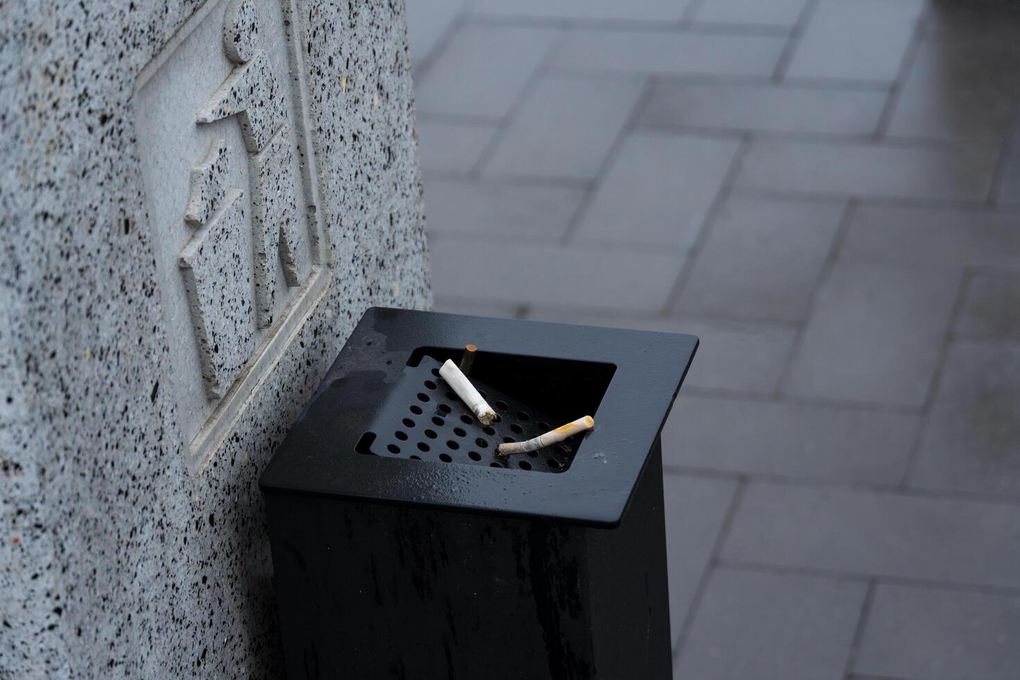 A black waste container filled with discarded cigarette butts stands on a wet pavement under gray skies in a bustling city photo