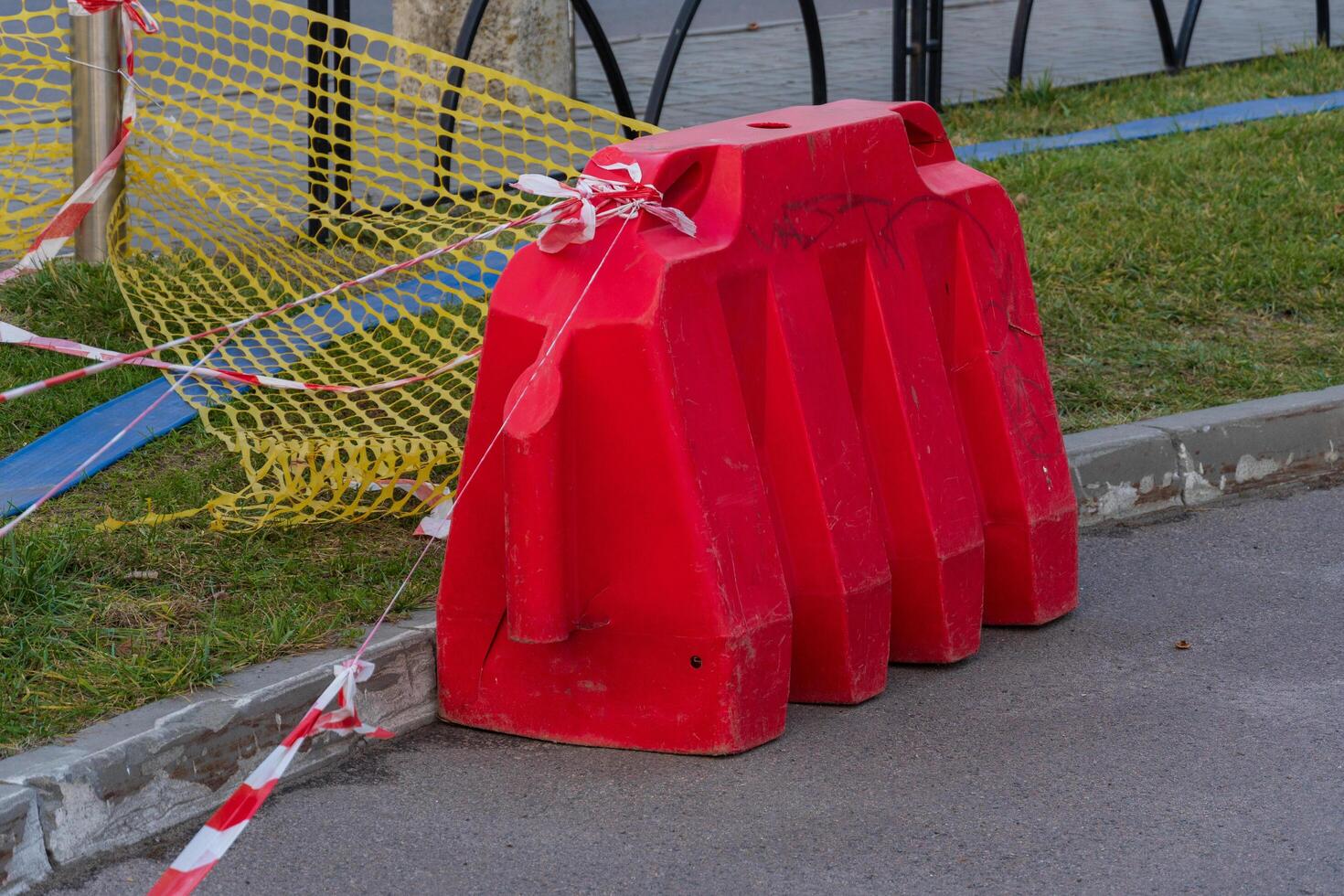 A vivid red plastic barrier stands prominently on the roadside, cordoned off with caution tape as workers prepare for upcoming construction. Sunlight illuminates the area, enhancing visibility photo
