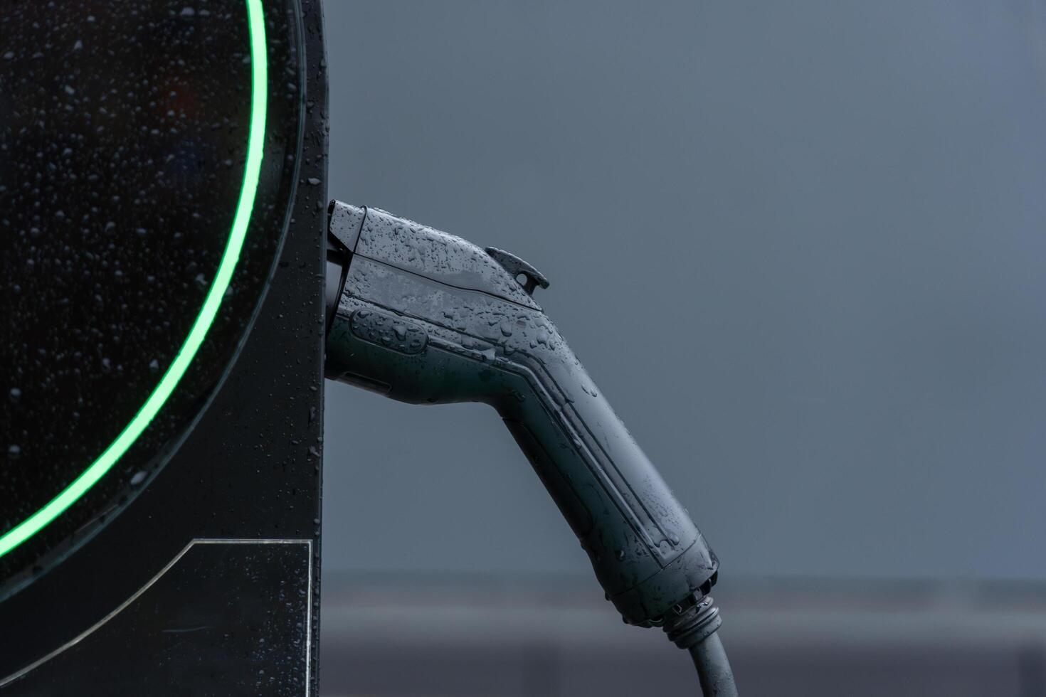 In a modern urban environment, an electric vehicle charging station stands out under the drizzle, showcasing a sleek charging cable ready for connection photo