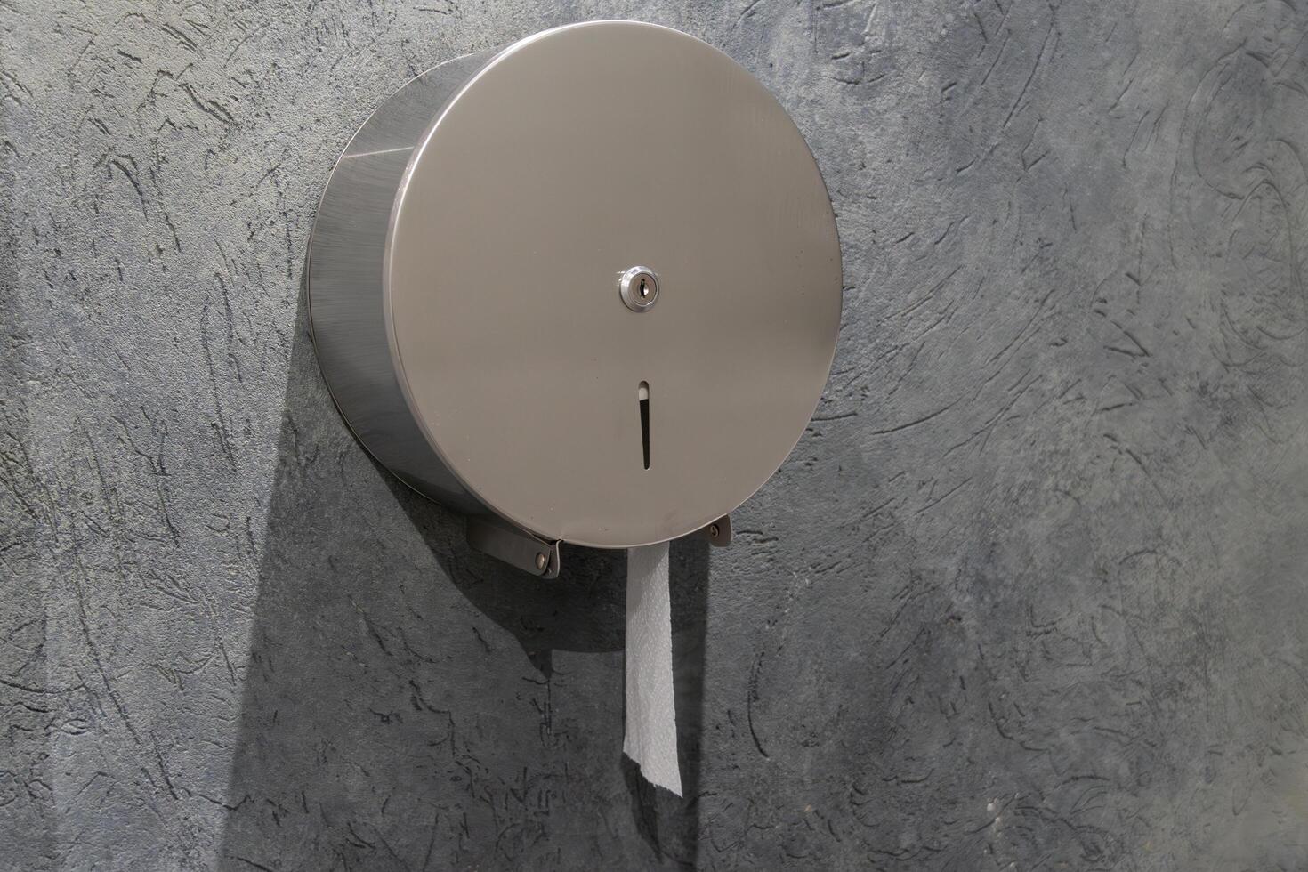 A round stainless steel toilet paper dispenser is securely mounted on a textured gray wall inside a public restroom. The dispenser has a small opening for easy paper access photo