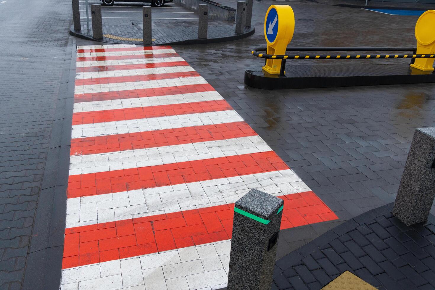 A vibrant red and white striped pedestrian crossing is seen at an urban intersection. Wet pavement reflects the colors as vehicles pass by, illustrating the activity of city life during a rainy day photo