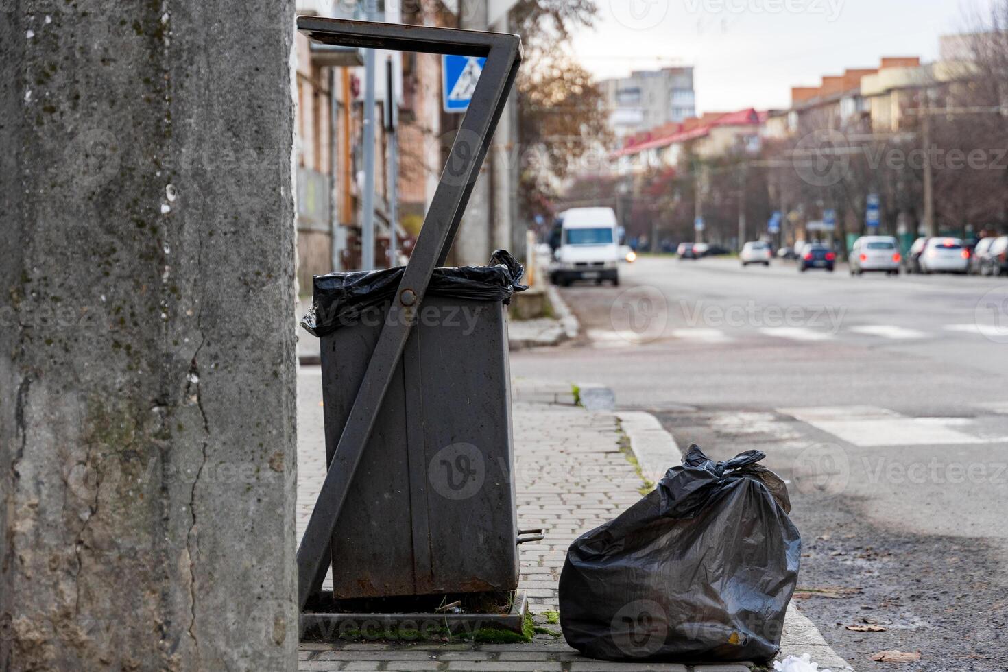 A grey trash container sits beside a sidewalk, partially filled with waste. A large black garbage bag lies on the ground nearby, emphasizing urban littering photo