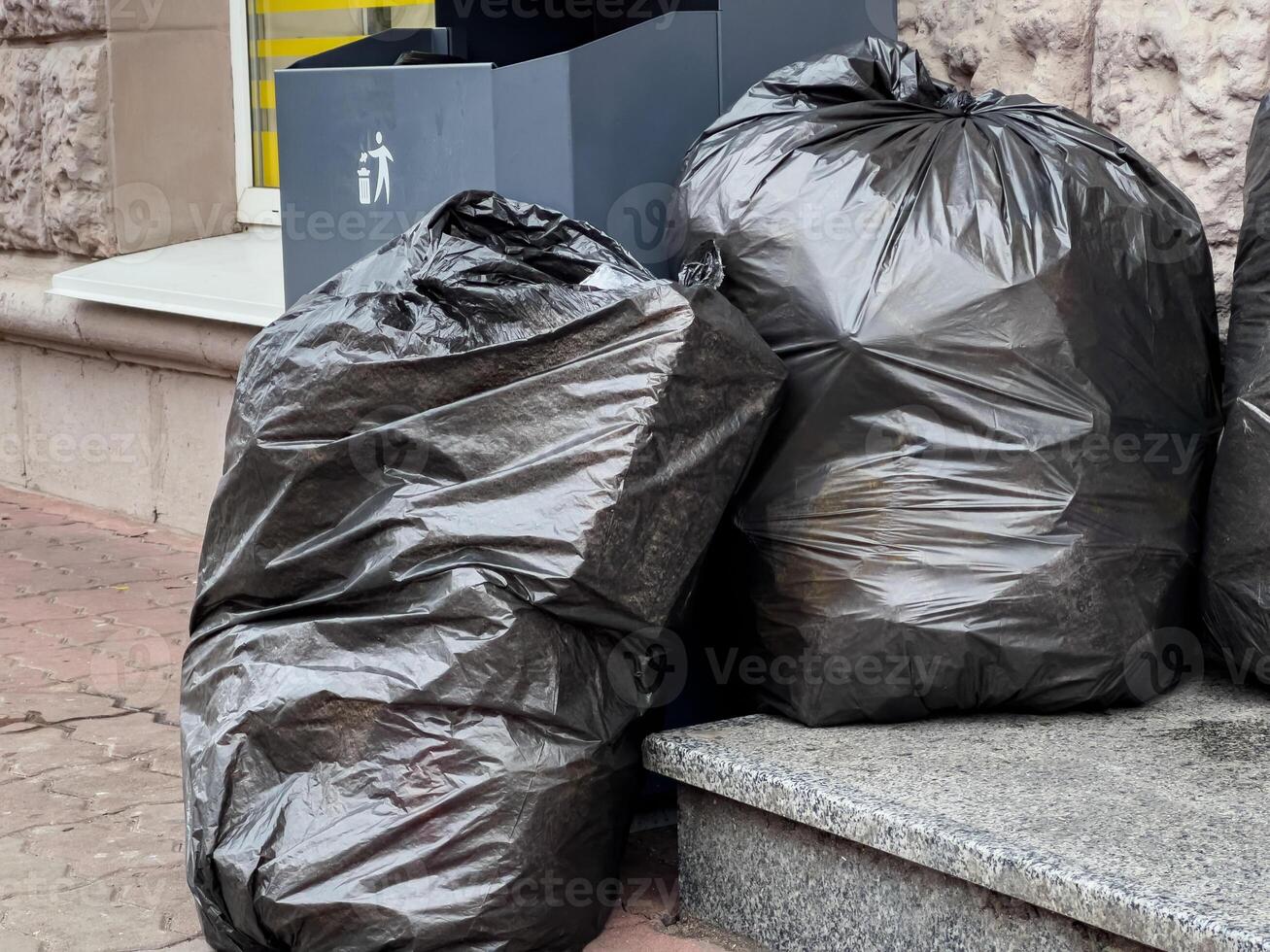 Black garbage bags lean against a clean, modern trash container on a city sidewalk. The sunlight highlights the need for environmental awareness and responsible waste management in urban areas photo