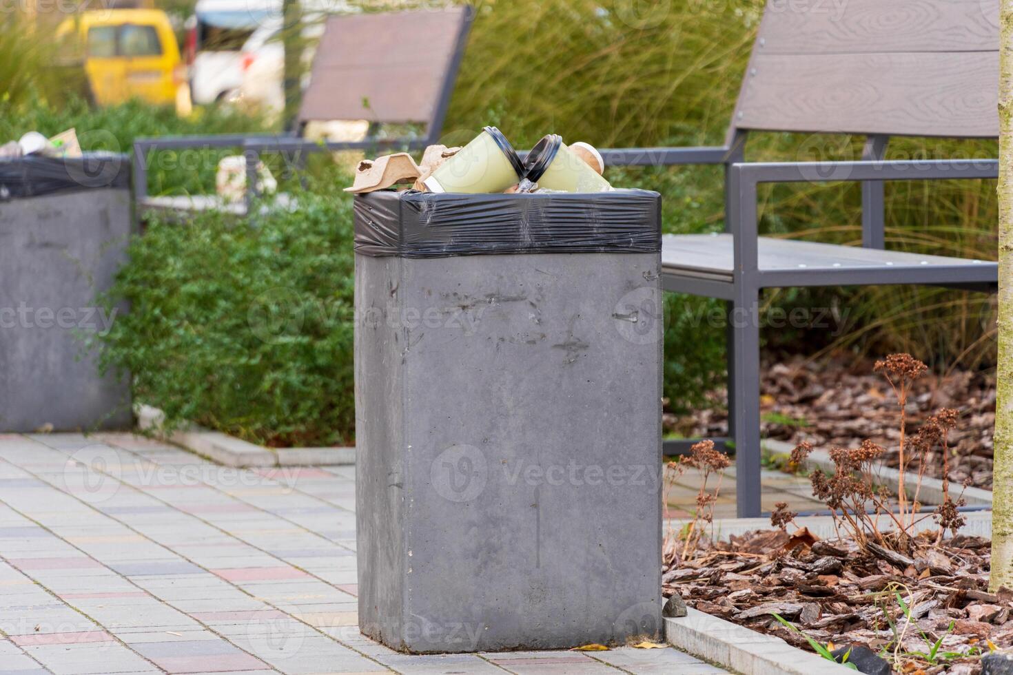 A concrete trash bin stands filled with waste bags, surrounded by greenery and benches in a bustling urban park. This spot reflects both convenience and environmental responsibility photo
