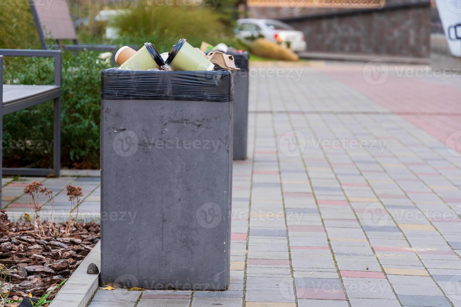 A concrete garbage bin filled to capacity with trash bags sits on a bustling city sidewalk, surrounded by plants and pavers under a clear sky. People pass by, highlighting urban waste challenges photo