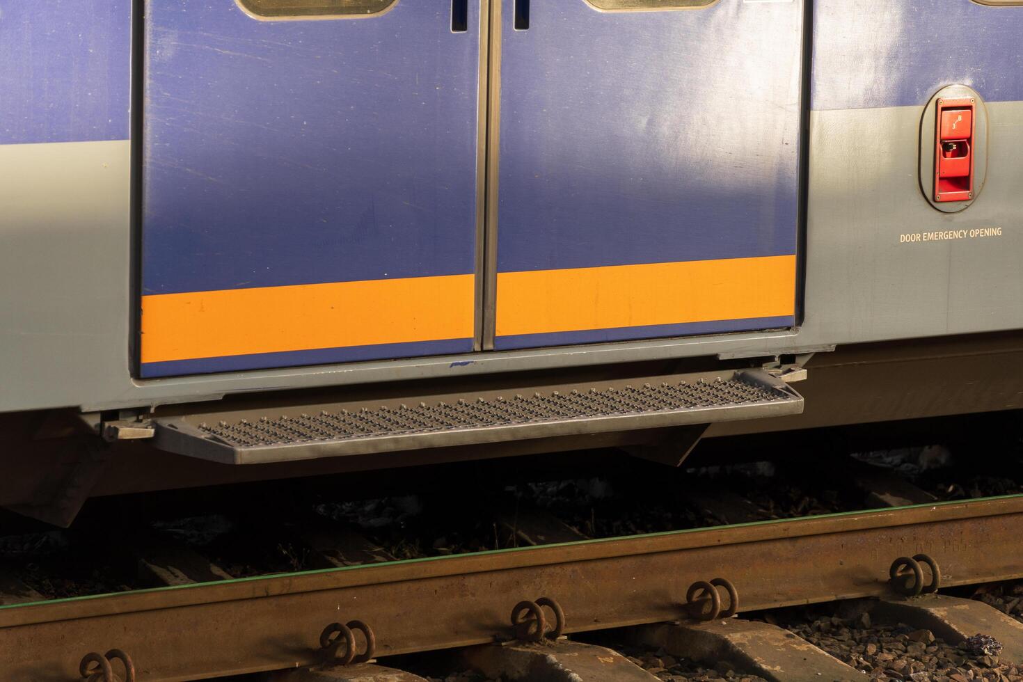 Close-up of a modern electric train door positioned at a railway station. The vibrant colors contrast with the rail tracks and surrounding environment during sunset, creating a striking visual photo