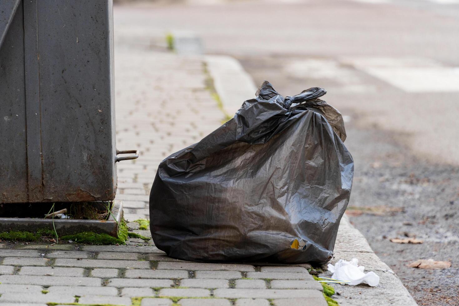 A black garbage bag is placed beside a recycling container on a city street. Litter can be seen on the pavement, highlighting issues of waste management in urban areas photo