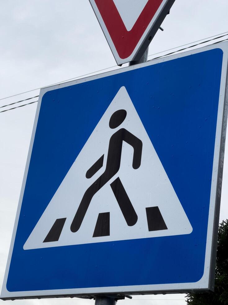 A triangular blue warning sign depicts a person walking amidst construction barriers, notifying drivers of pedestrian presence in a work zone. This sign is positioned at a busy intersection photo