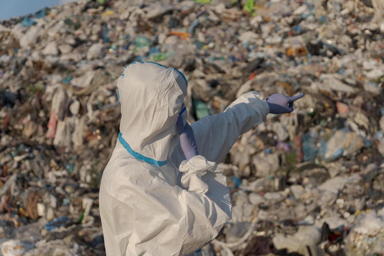 A sanitation worker in protective clothing gestures towards an expansive landfill brimming with plastic waste and debris photo