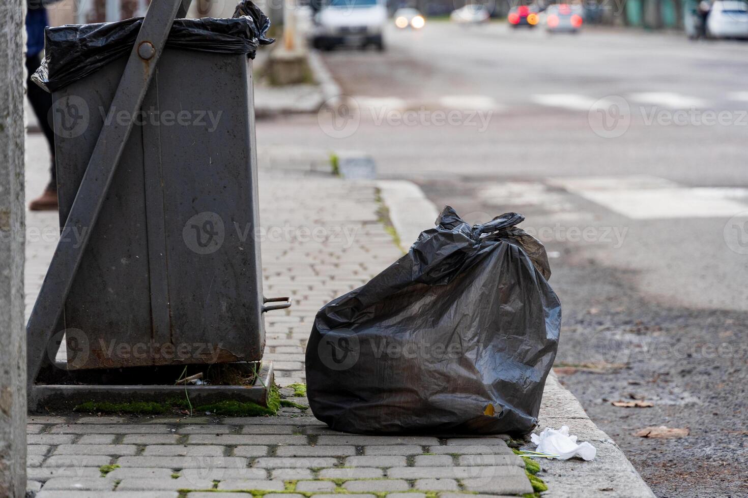 A garbage container stands near the curb of an urban street. Black trash bags are placed next to the bin, highlighting the issue of waste management and recycling photo