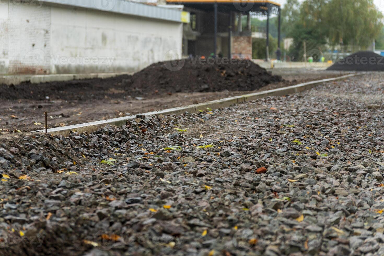 Gravel and dirt are spread along the sidewalk of an urban road in the midst of construction. Industrial buildings are present in the background as work continues photo