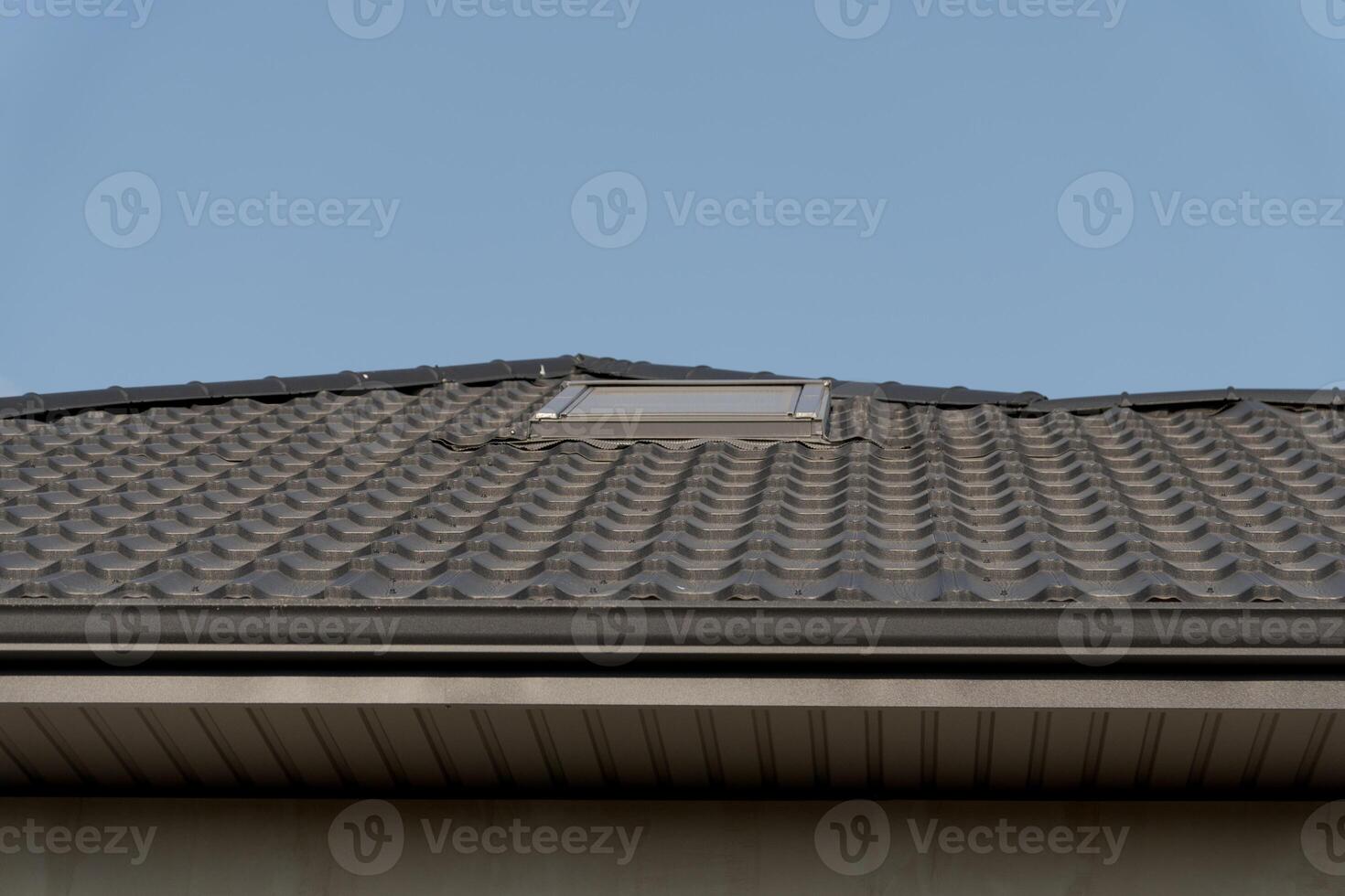 A modern residential roof features sleek metallic tiles with a skylight. The rooftop design blends functionality and aesthetics against a clear blue sky photo