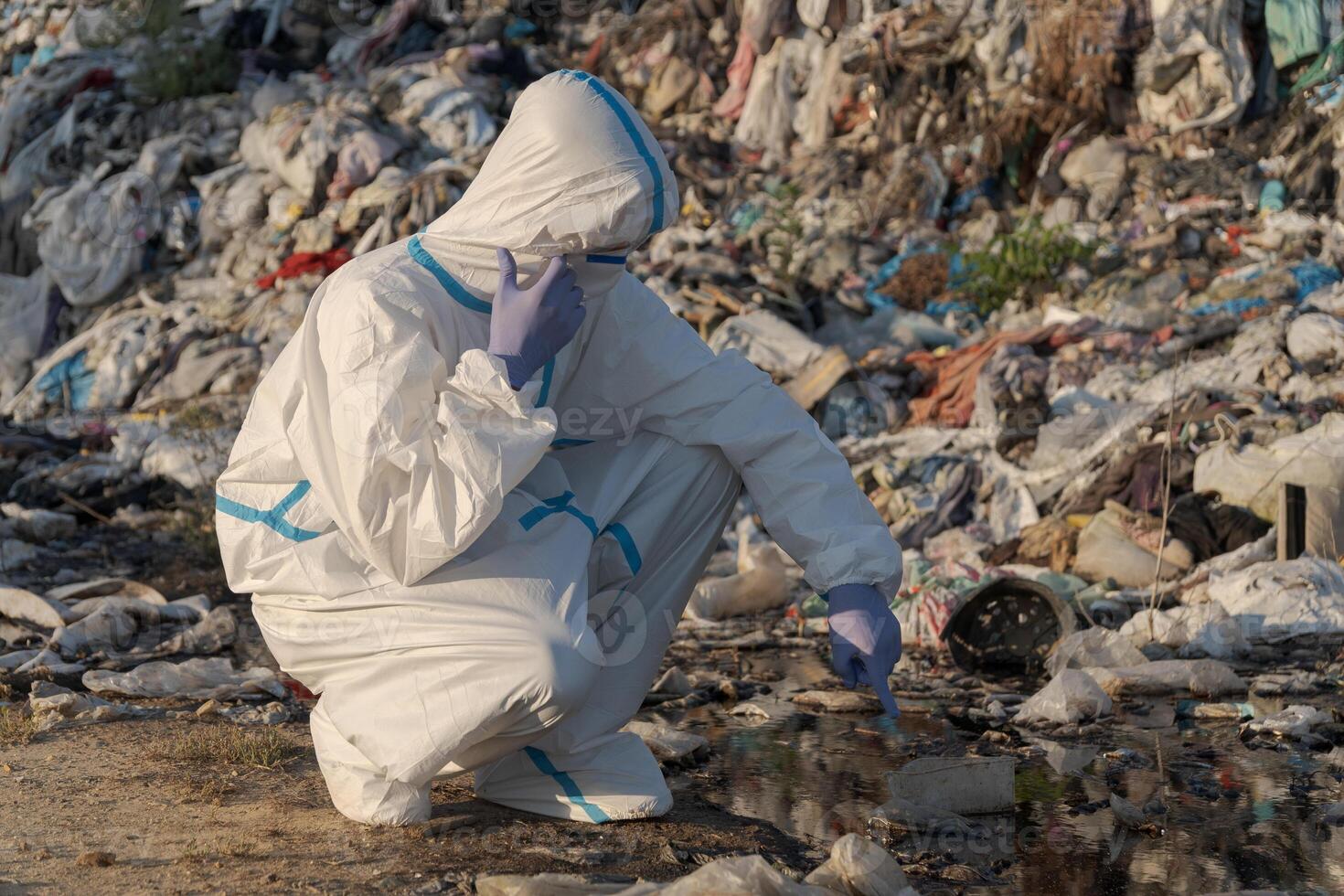 A person dressed in protective gear examines polluted water at a landfill filled with trash and plastic waste photo