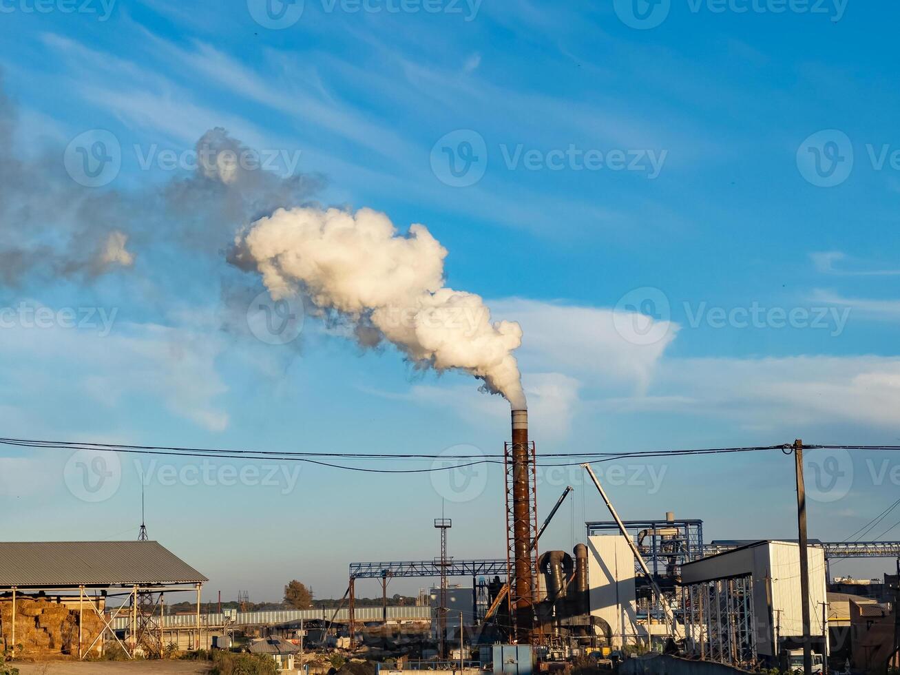 Production activities generate thick smoke from the factory's smokestacks, impacting the surrounding environment while machinery operates under a bright sky photo