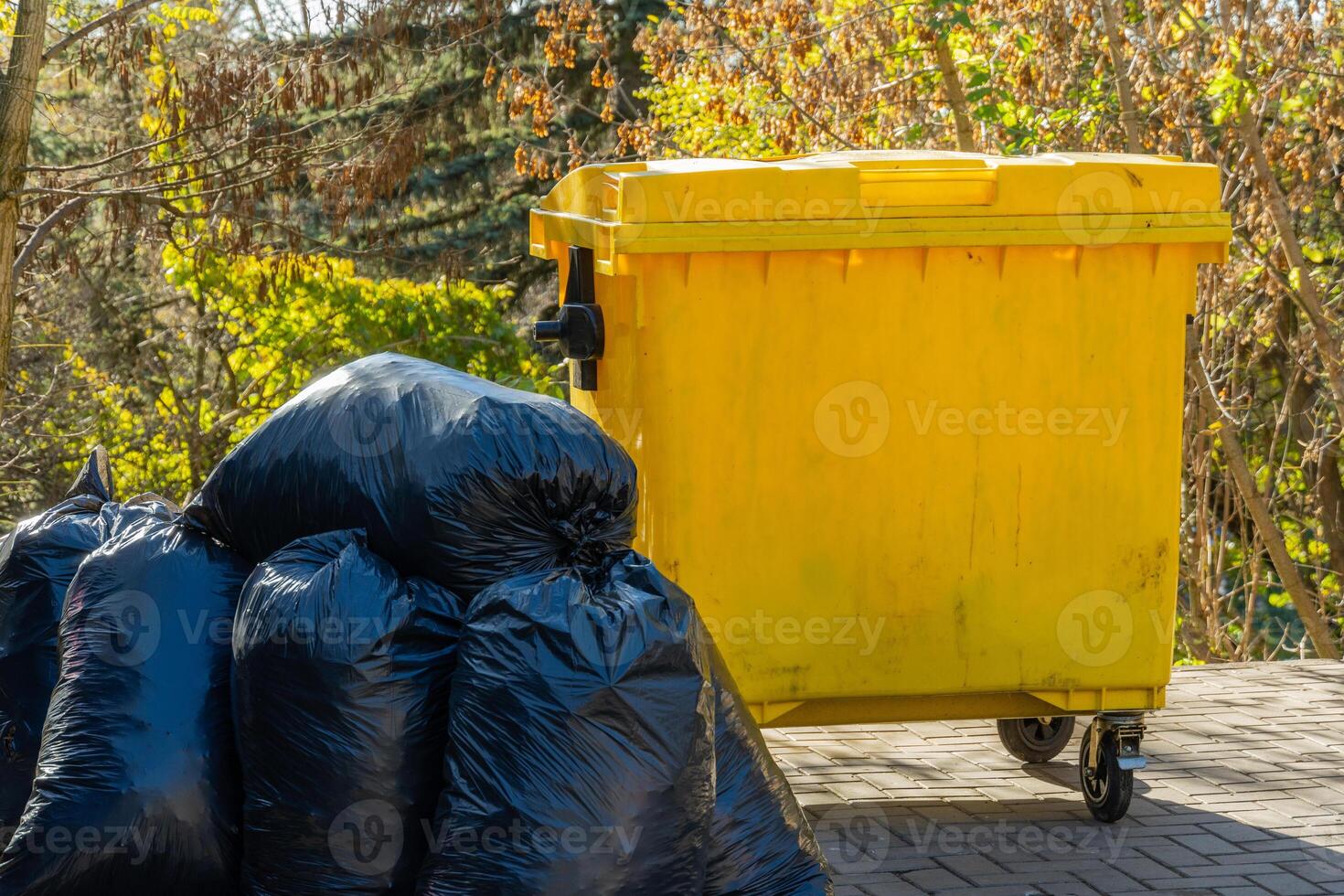 Several black garbage bags are piled next to a yellow waste container in a residential outdoor setting. The greenery around adds to the environmental context photo