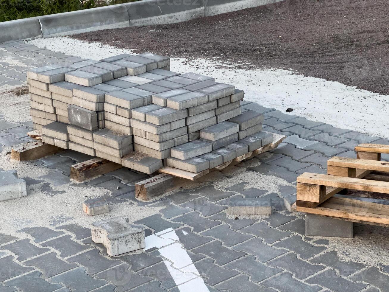 Sturdy concrete pavers are stacked on wooden pallets, surrounded by loose sand and gravel at an urban construction site near a paved street, ready for installation photo